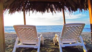 Two white lounge chairs under a thatched roof, facing a beach with clear blue water. - Exclusive Ocean View Apartment - Reserva del Mar (Santa Marta)
