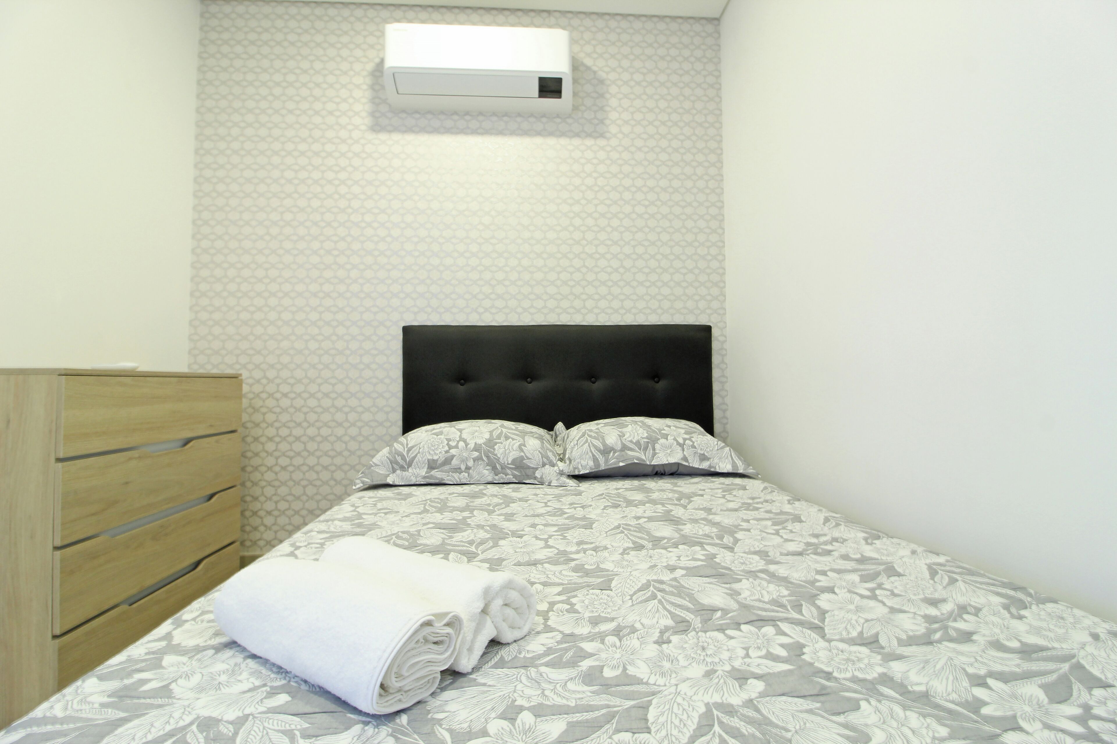 Ein ordentlich gemachtes Bett mit einem blumigen Überwurf, einem schwarzen, mit Stoff bezogenen Kopfteil und einer Holzkommode mit Schubladen.