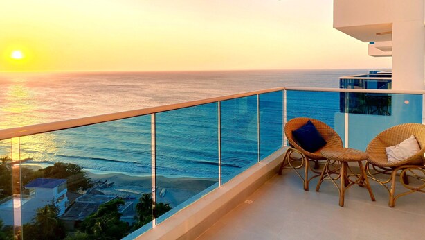A balcony with a view of the ocean at sunset, wicker chairs, and a glass railing. - Exclusive Ocean View Apartment - Reserva del Mar (Santa Marta)