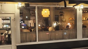 Un restaurante con una zona de bar visible a través de una ventana, que presenta una lámpara colgante y un letrero que dice 'DA DANTEL'.