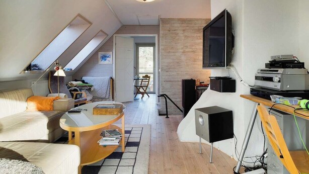 Un moderno loft con techo alto, techo inclinado con tragaluz, televisor de pantalla plana grande montado en la pared, mesa de comedor de madera, sofá y mesa de centro.