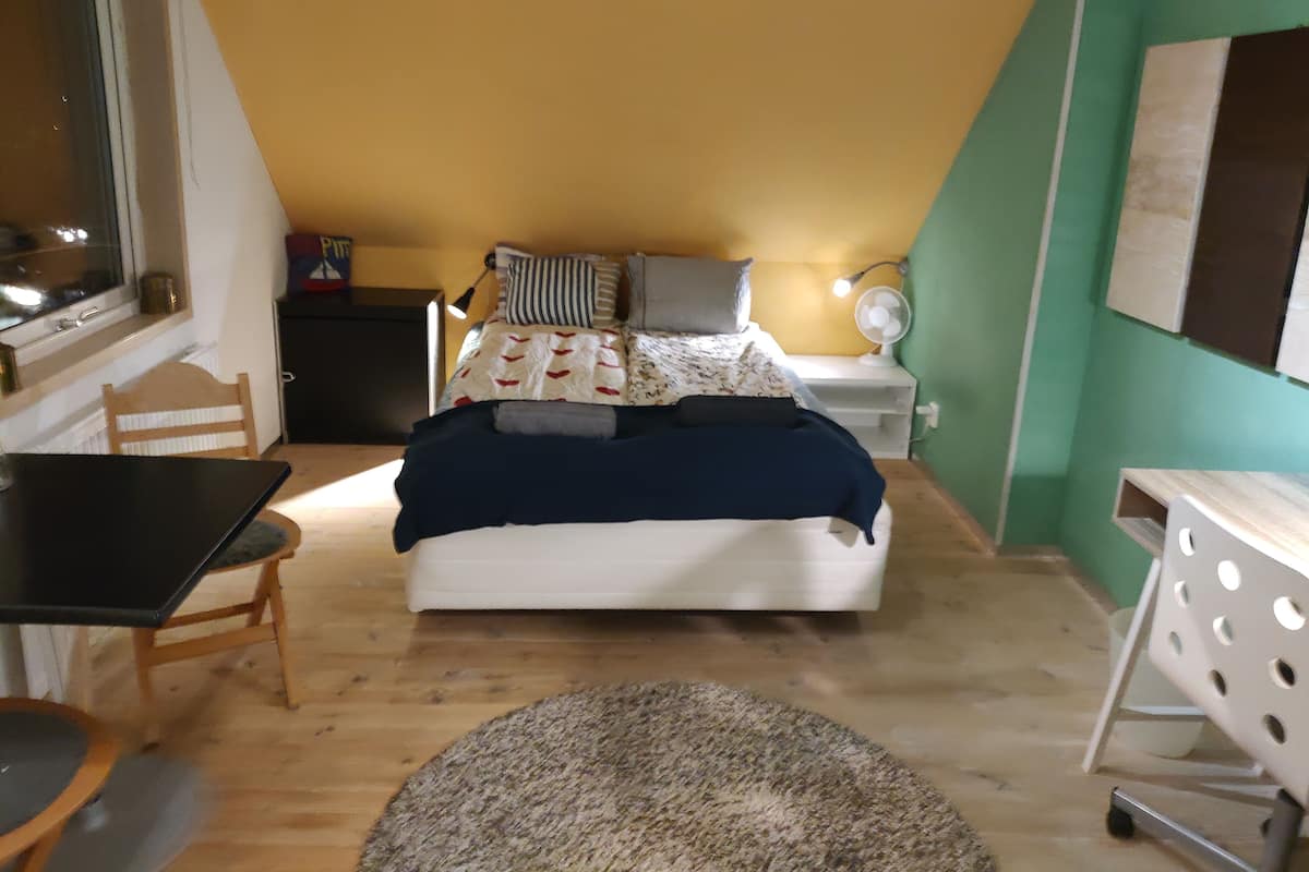 A bedroom with a bed, bedside table, lamp, and a small desk with a chair.