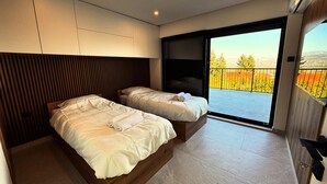 Un dormitorio moderno con dos camas, una puerta corredera a un balcón y vistas del follaje otoñal.
