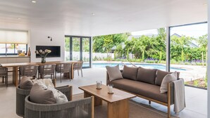 Una sala de estar moderna con un sofá, sillas, una mesa de centro y una televisión. Se ve una ventana grande con vista a una piscina y vegetación.