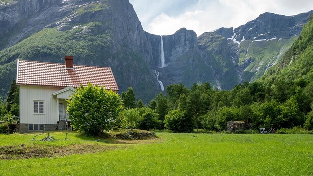 Une maison avec un toit recouvert de tuiles rouges, entourée de champs verdoyants et avec des montagnes en arrière-plan.