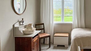 Une chambre à coucher comprenant un lit, une commode en bois, une chaise, un petit banc, un miroir et une fenêtre avec des rideaux.