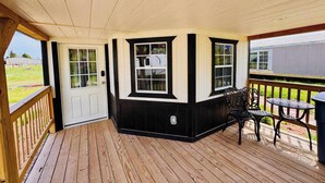 En inglasad veranda med trägolv, en vit dörr och svarta lister. Det finns två fönster med vita karmar, ett litet bord med stolar och en metallbehållare för sopor.