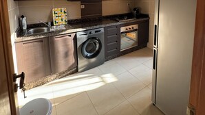 A compact kitchen with dark cabinets, a stainless steel washing machine, a built-in oven, and a refrigerator.