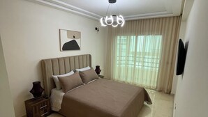 A bedroom with a large bed, a bedside table, a lamp, a picture on the wall, and a window with curtains.