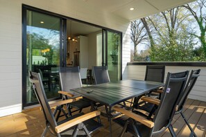 Eine Terrasse im Freien mit Tisch und Stühlen, eine Glasschiebetür zu einer Küche und im Hintergrund Bäume sowie ein Gebäude.