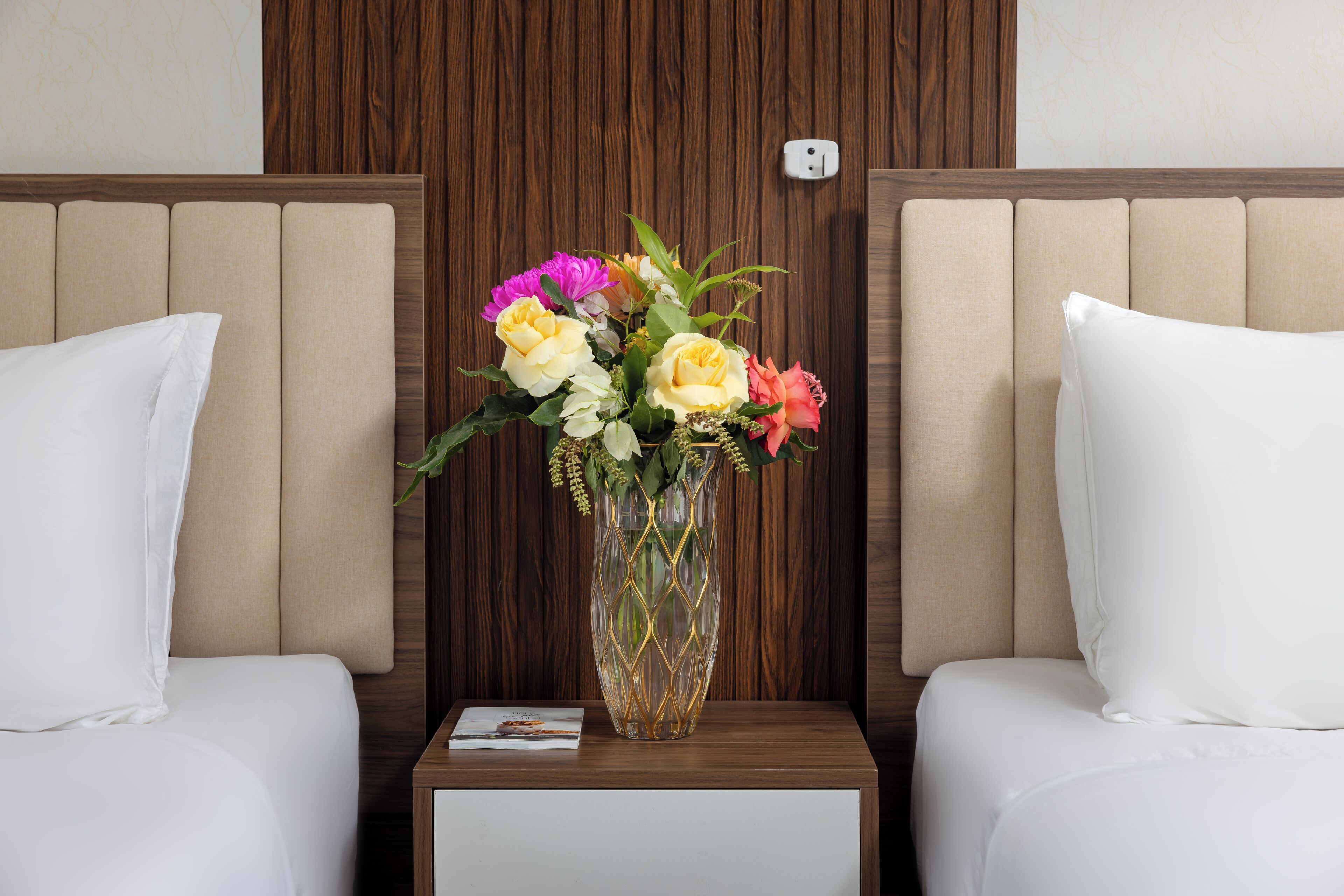 A hotel room with two beds, a wooden headboard, a nightstand with a vase of flowers, and a small table with a book.