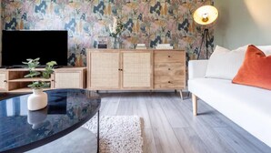 A modern living room with a flat-screen TV, a wooden cabinet, a white sofa with an orange pillow, and a floral wallpaper. - Charming Whitehurst Drive Nest in Manchester (Manchester)