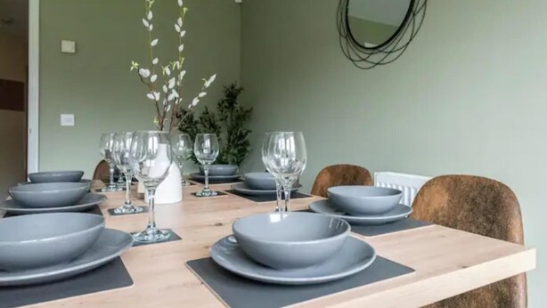 A dining table set with grey plates and wine glasses, a vase with white flowers, and a mirror on the wall. - Charming Whitehurst Drive Nest in Manchester (Manchester)