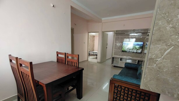 A dining area with a wooden table and chairs, a sofa, and a television in the background. - Ramamurthy/sleeps6/2bedroom/tv/wifi (Bengaluru)