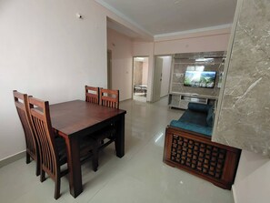 A dining area with a wooden table and chairs, a sofa, and a television in the background. - Ramamurthy/sleeps6/2bedroom/tv/wifi (Bengaluru)