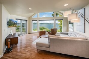 A spacious living room with a large window overlooking a body of water, a modern sofa, a wooden coffee table, and a television mounted on the wall. - Waterfront Home with Game Room, Fireplaces, Kayaks, AC and Private Beach Access (Camano)