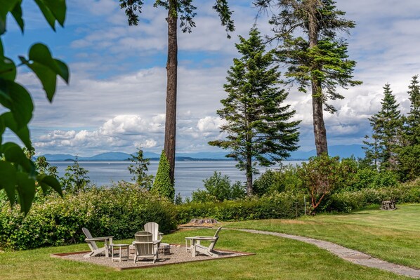 A serene lakeside setting with a patio area featuring Adirondack chairs and a table, surrounded by tall trees and a view of the lake and distant mountains.