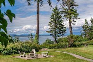 Un entorno tranquilo junto al lago, con un patio que incluye sillas Adirondack y una mesa, rodeado de altos árboles y vistas al lago y montañas distantes.
