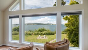 A room with large windows overlooking a grassy area and water, with two brown chairs in the foreground. - Waterfront Home with Game Room, Fireplaces, Kayaks, AC and Private Beach Access (Camano)