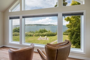 Una habitación con ventanas amplias que dan a un área verde y al agua, con dos sillas marrones en primer plano.