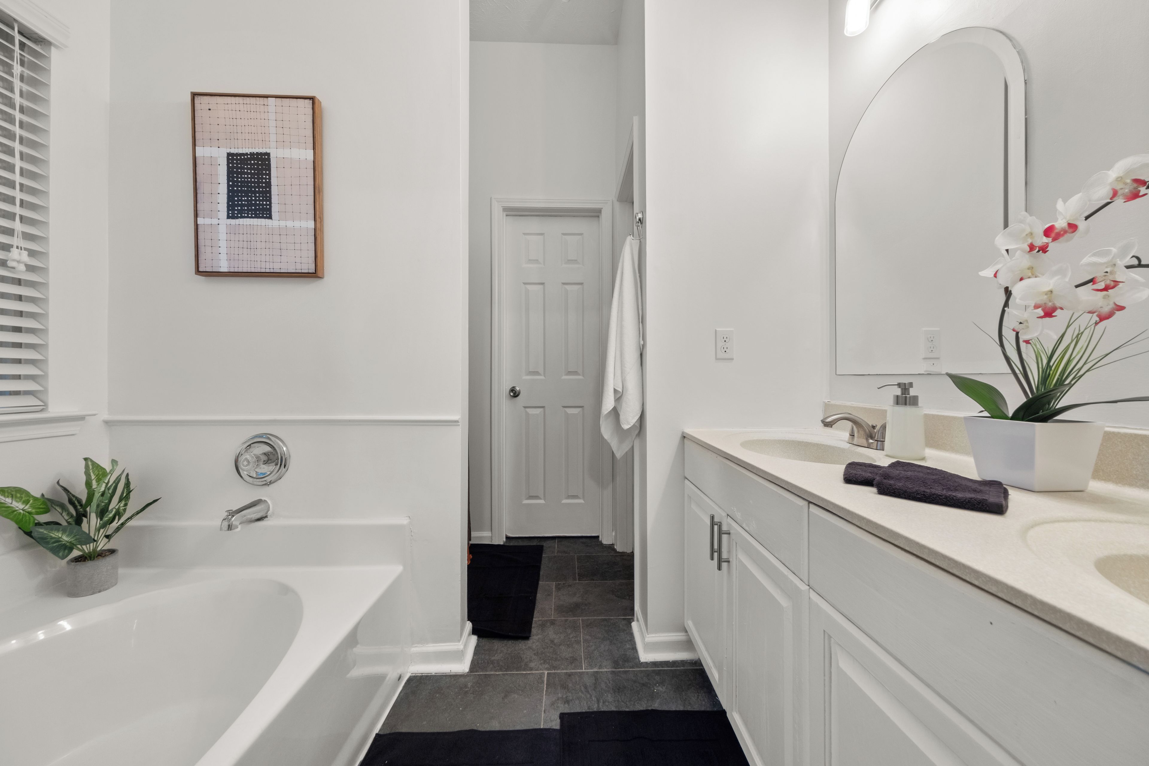 Bathroom 1: Primary en-suite: double vanity, arched mirror, white orchid arrangement, deep soaking tub, dark slate tile, framed art. Everything a full Atlanta group gathering weekend needs feels quietly handled in here.