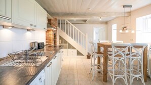 Una cocina con armarios blancos, encimera de madera y horno de ladrillo empotrado. Se ve una escalera que sube a un comedor con sillas de mimbre blancas y una mesa de madera.