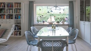 Un comedor con mesa de vidrio y sillas de metal, una estantería y una ventana que da a vistas de vegetación y un velero.