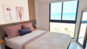 Un dormitorio moderno con una cama grande, una mesita de noche y una ventana con vistas a la playa.