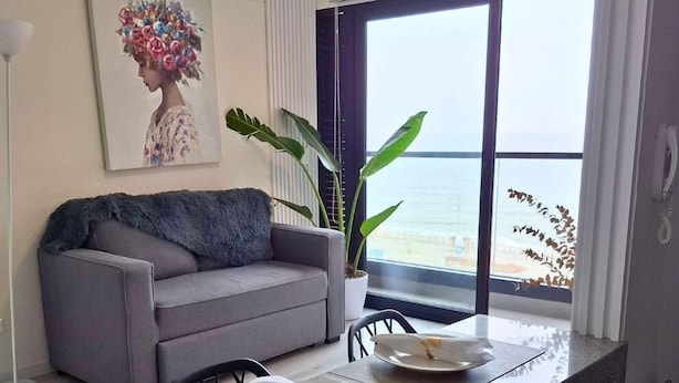 Una sala de estar moderna con un sofá gris, una planta grande en maceta, un cuadro y vista al mar a través de un ventanal amplio.