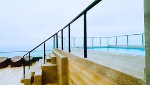 Zona junto a la piscina con escalones que bajan a ella, barandilla de cristal y vista al mar.
