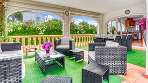 A covered outdoor patio with wicker furniture, a dining table, and a view of greenery and a building.