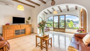 A spacious living area with a wooden TV stand, a sofa, a small table, and a large arched window offering a view of greenery.