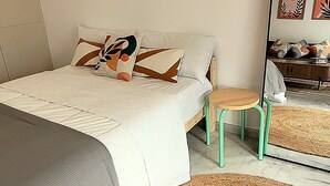 A bedroom with a bed, a small stool, a large woven rug, and a mirror reflecting the room.