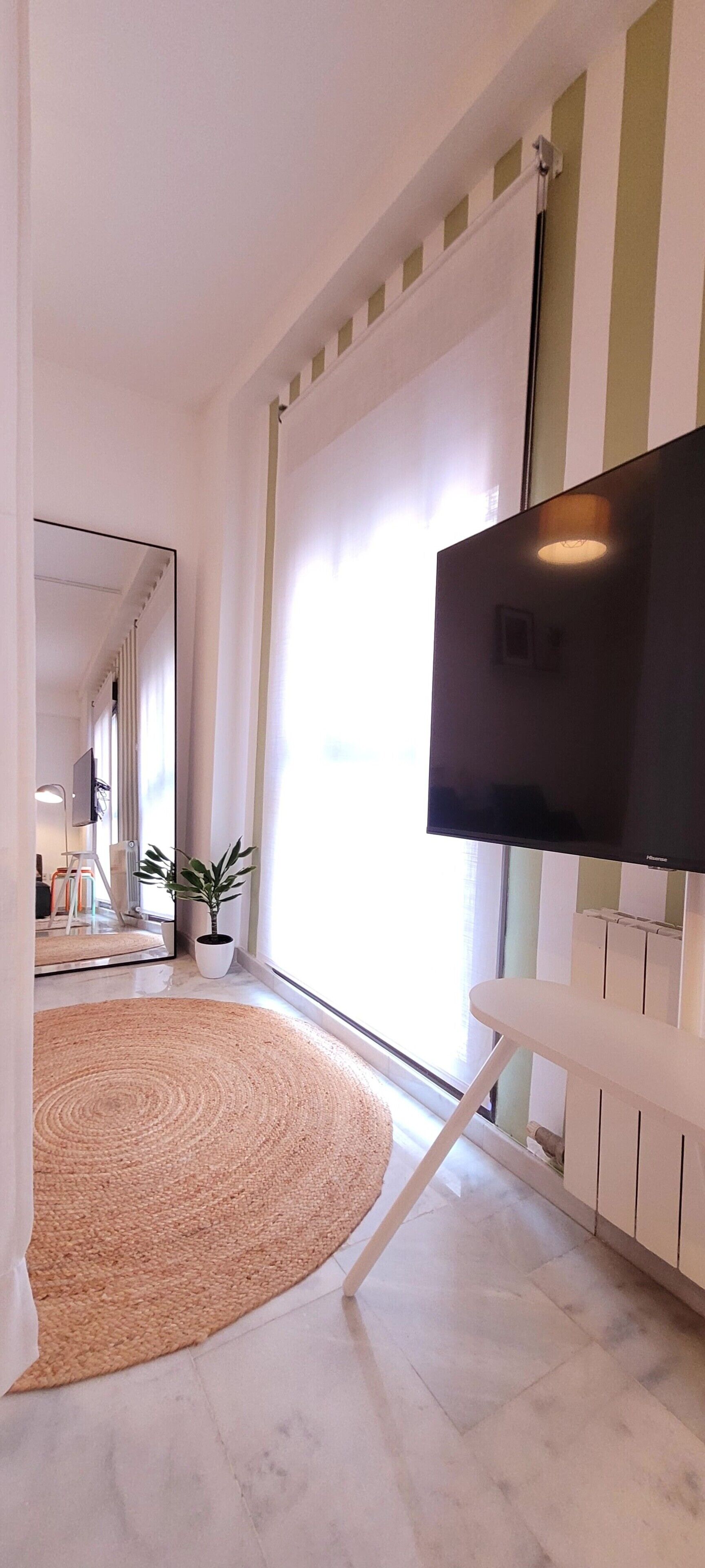 Un salon moderne avec un téléviseur à écran plat fixé sur un meuble, une grande fenêtre avec des rideaux, un tapis tissé et une plante en pot.