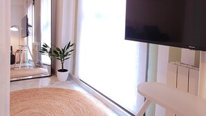 A modern living room with a flat-screen TV mounted on a stand, a large window with curtains, a woven rug, and a potted plant.