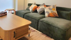 A living room with a green sofa, a wooden coffee table, a small round table, and a floor lamp. There are two framed artworks on the wall.