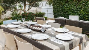 Un área de comedor al aire libre con una mesa lista para cuatro personas, rodeada de muebles de mimbre y un seto.