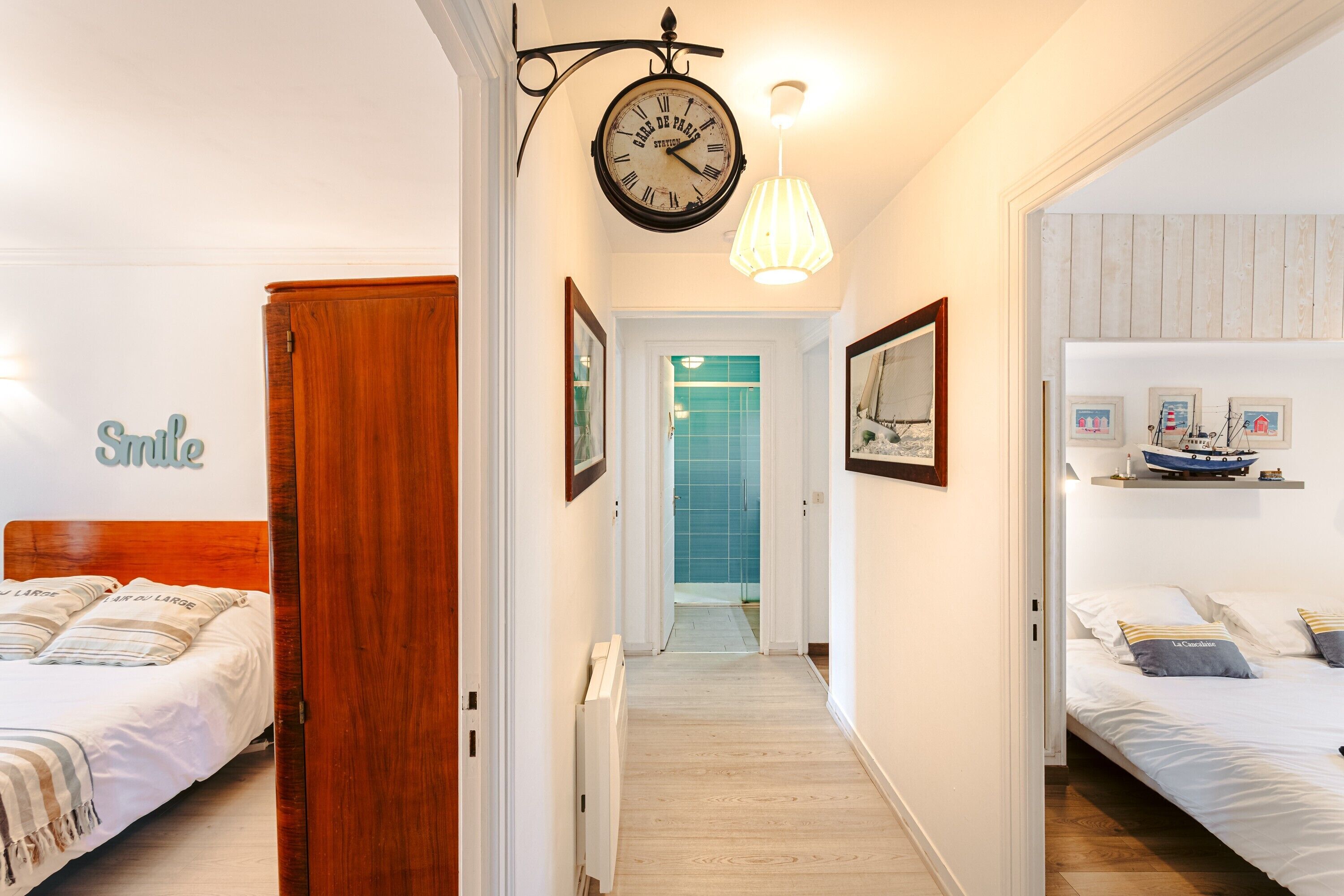 Een hotelkamer met een bed, een houten garderobe, een gang naar een badkamer, en een muur met een klok en afbeeldingen in lijsten.