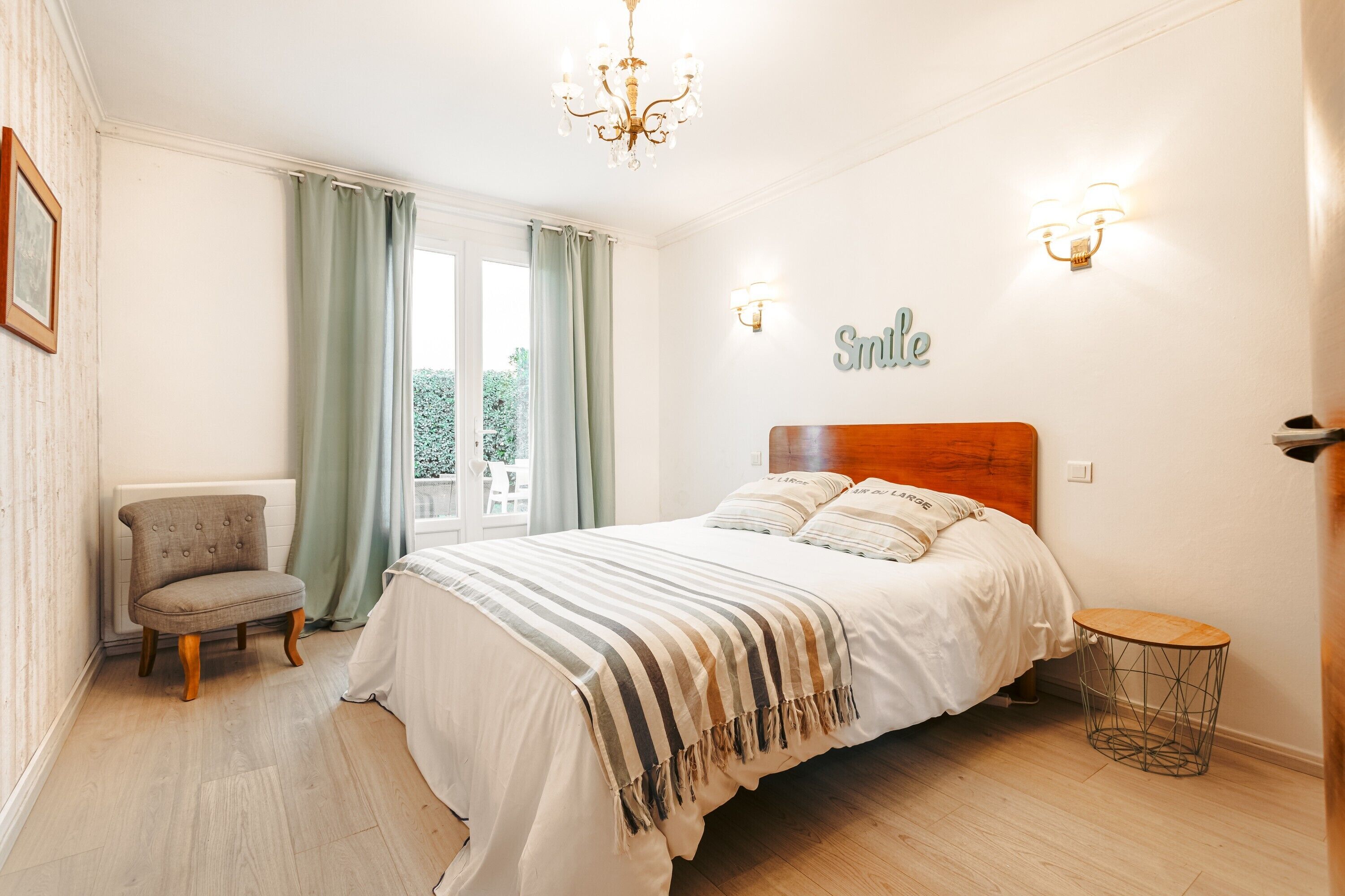 Une chambre à coucher comprenant un lit, une chaise, une petite table ronde, un lustre et une fenêtre avec des rideaux.