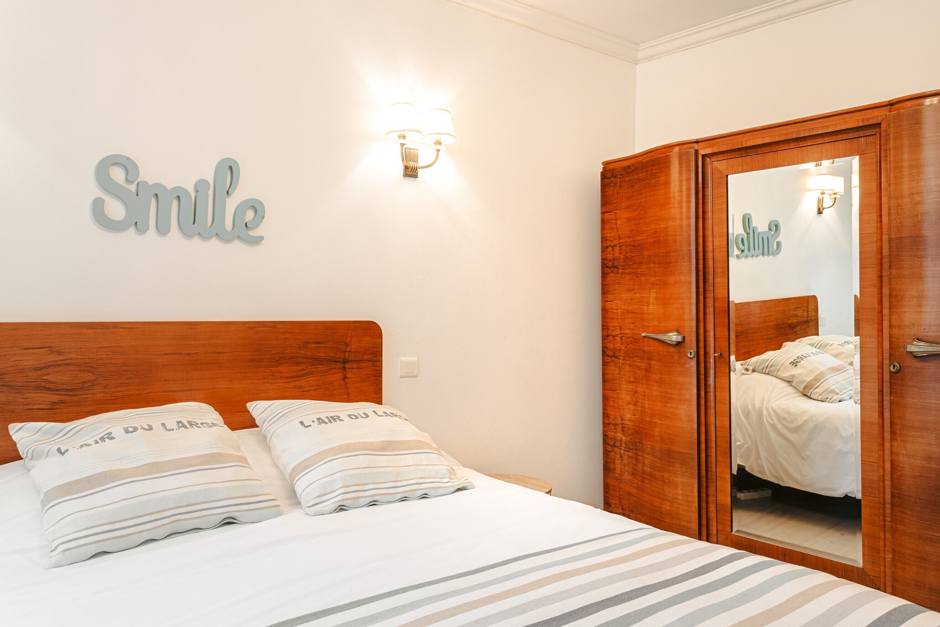 Une chambre avec une tête de lit en bois, un lit avec une literie à rayures, deux oreillers ornés de motifs, une lampe fixée au mur et une armoire avec un miroir.