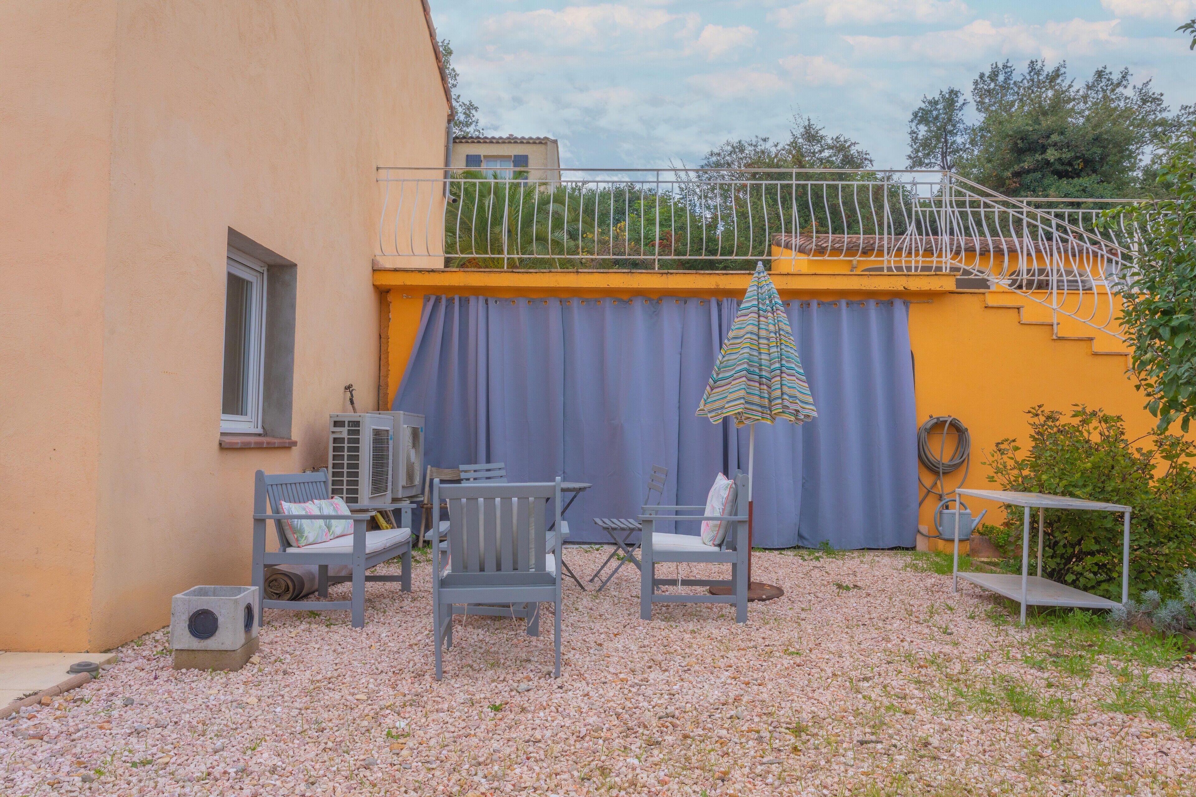 Un espacio al aire libre con sillas y una mesa, un porche cubierto y una escalera que lleva a un nivel superior.