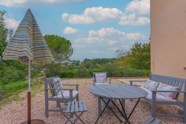 An outdoor seating area with a table and chairs, a striped umbrella, and a view of greenery and hills.