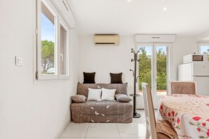 A bedroom with a sofa bed, a dining table, a chair, an air conditioning unit, and a view of trees through the window.