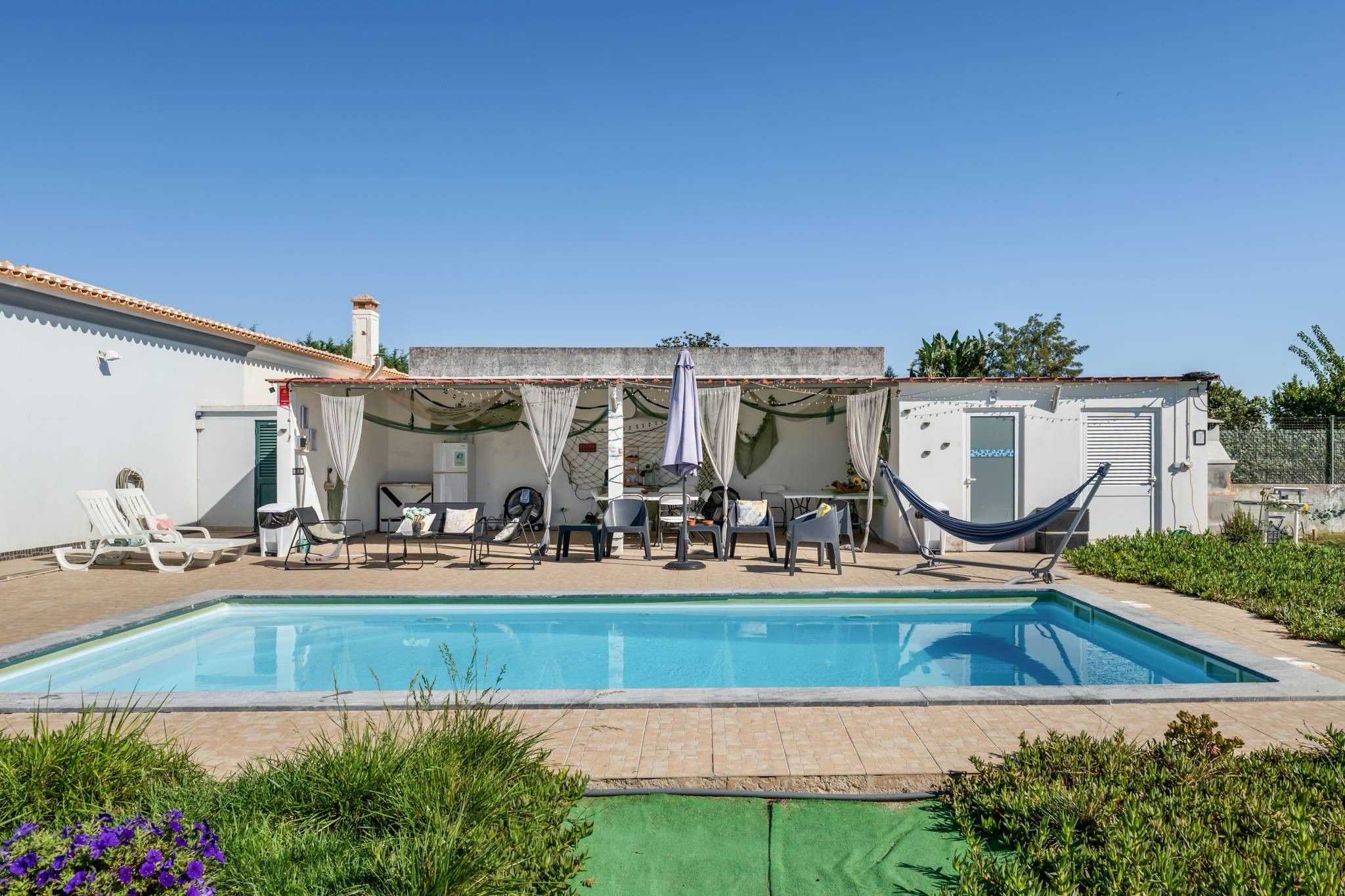 Una piscina con sillones de descanso y una hamaca, rodeada de vegetación y un edificio con techo de tejas.