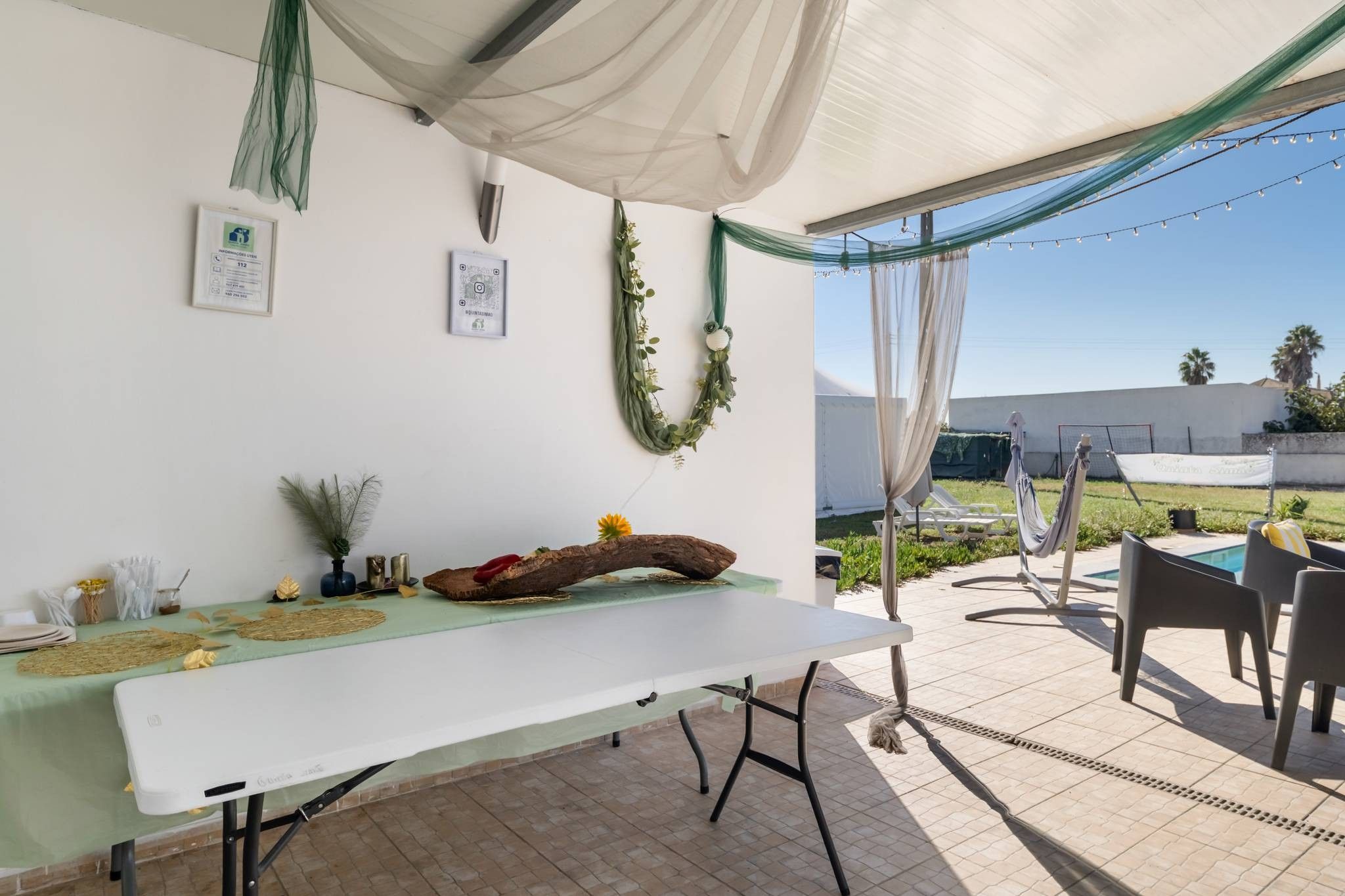 An outdoor patio with a table set for dining, surrounded by chairs, a pool, and a covered area with draped fabric.