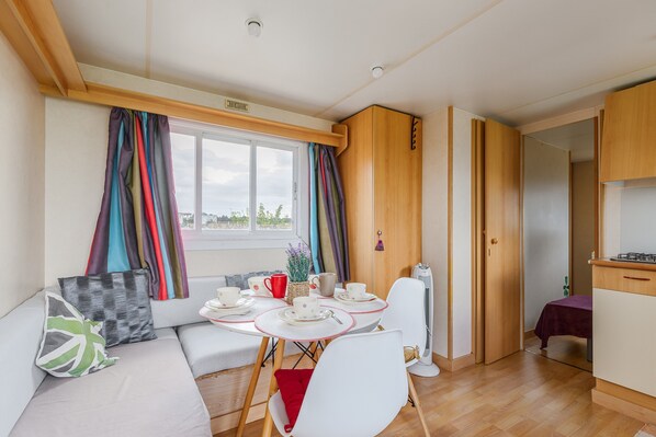 A compact room with a dining table, chairs, a sofa, a small kitchen area, and a window with curtains.