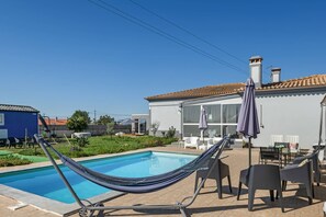 Piscina com rede e espreguiçadeiras, um edifício branco com telhado de azulejos e céu azul límpido.