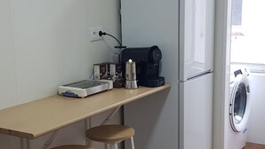 Een compacte keuken met een witte koelkast, een koffieapparaat en een wasmachine. Er staan twee krukken bij een klein tafeltje.