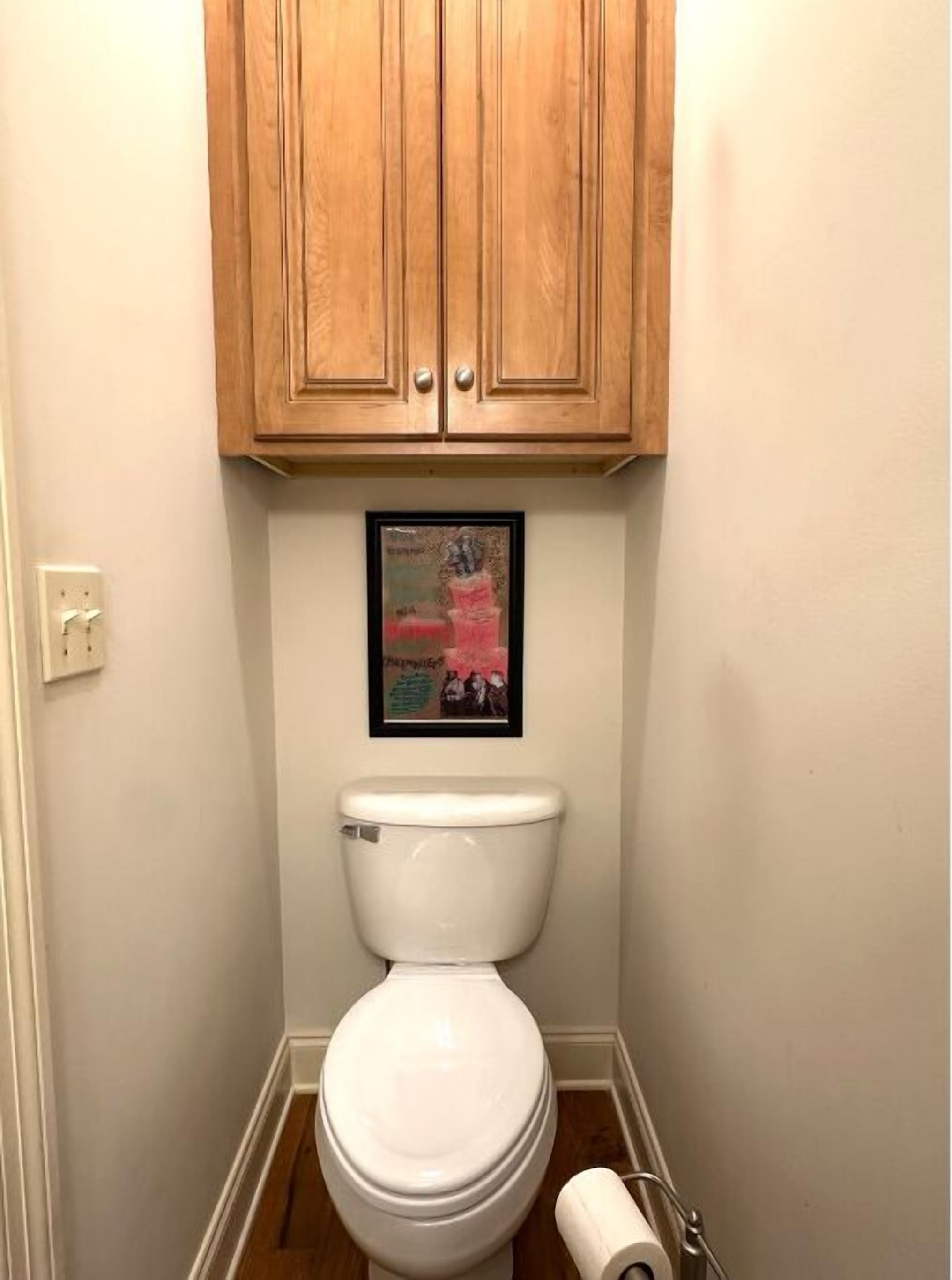 Une salle de bain avec des toilettes blanches, des meubles en bois au-dessus et un tableau encadré au mur.