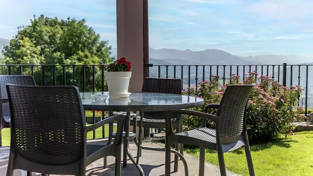 Une table ronde en verre, quatre chaises, un vase blanc avec des fleurs rouges et une vue sur les montagnes.